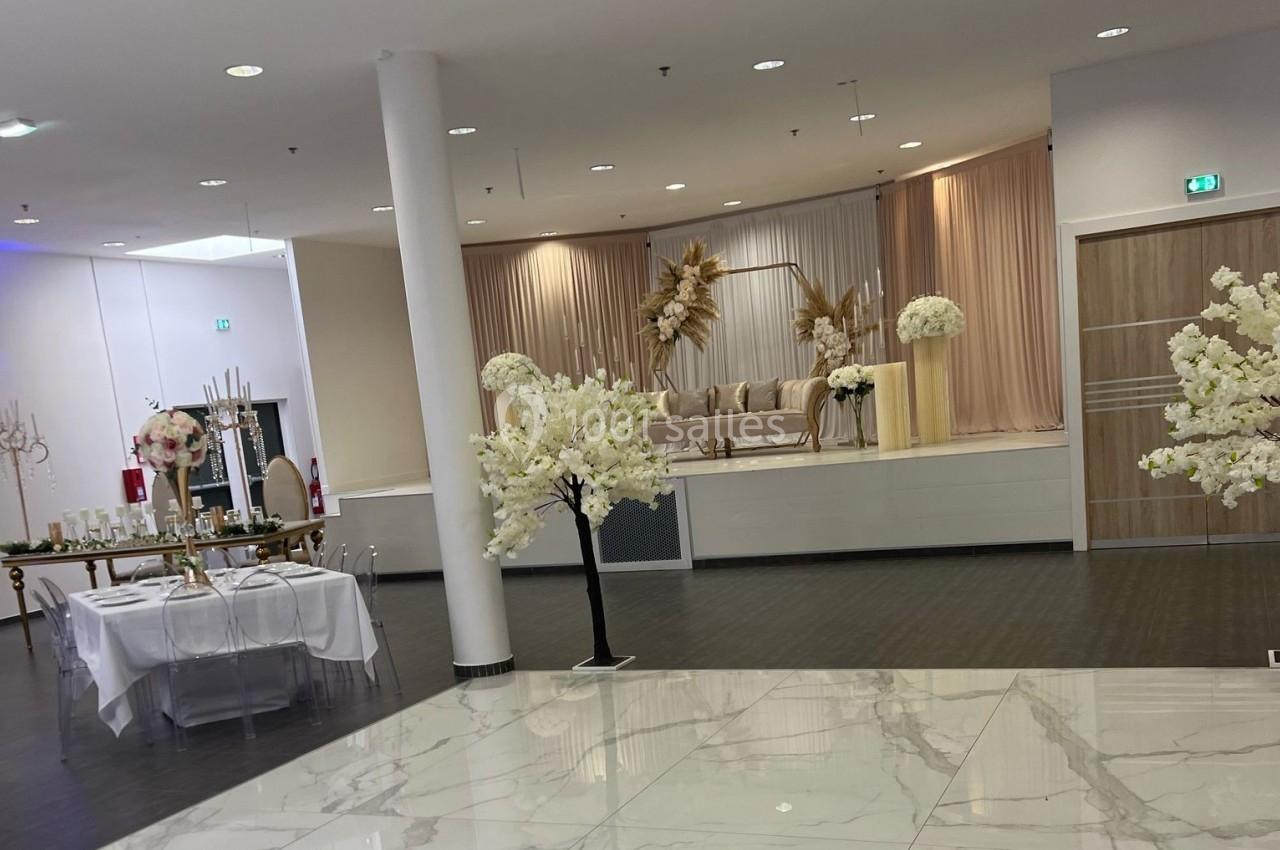 Salle de réception élégante avec scène décorée de fleurs blanches et beiges, tables dressées et sol en marbre.