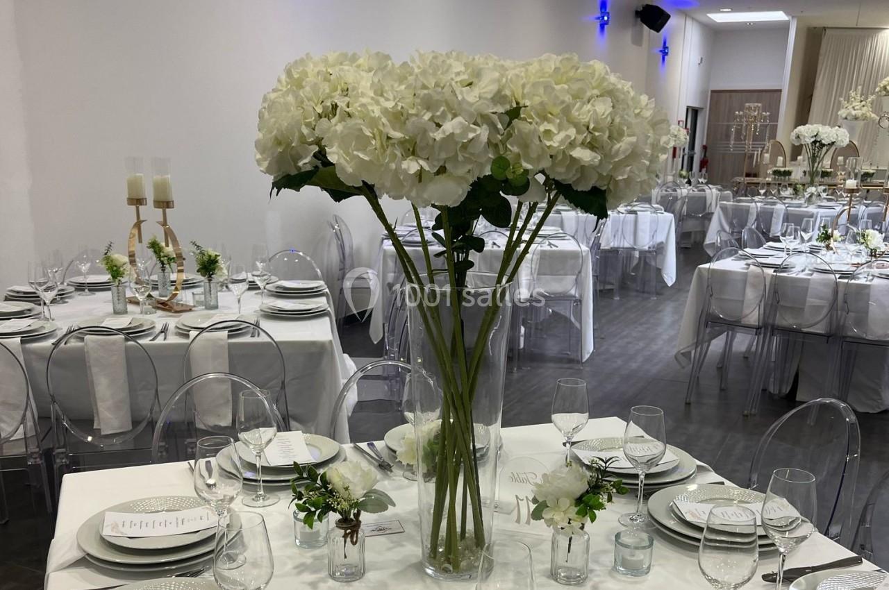 Salle de réception décorée avec des tables dressées, des chaises transparentes et un grand bouquet d'hortensias blancs.