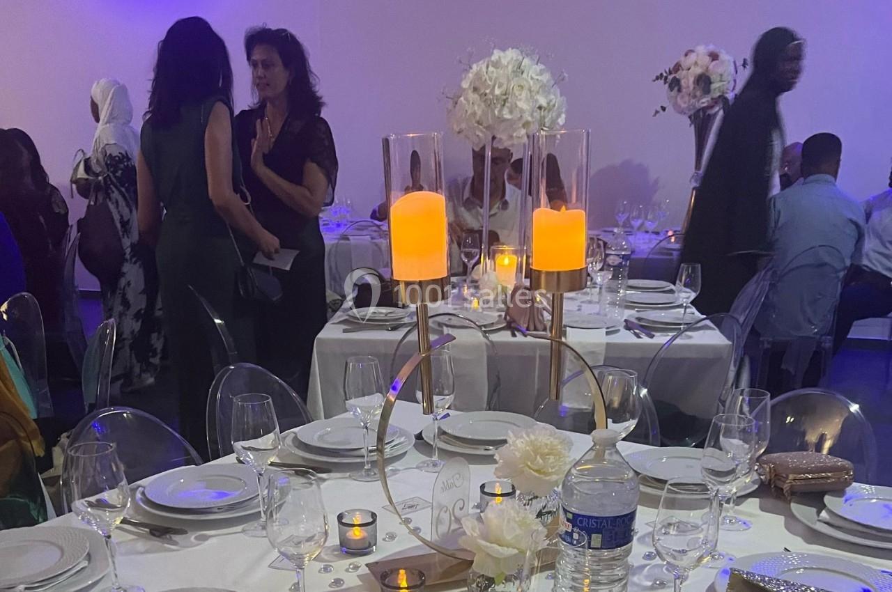 Salle de réception décorée avec des tables dressées, des bougies allumées et des fleurs blanches, invités en arrière-plan.