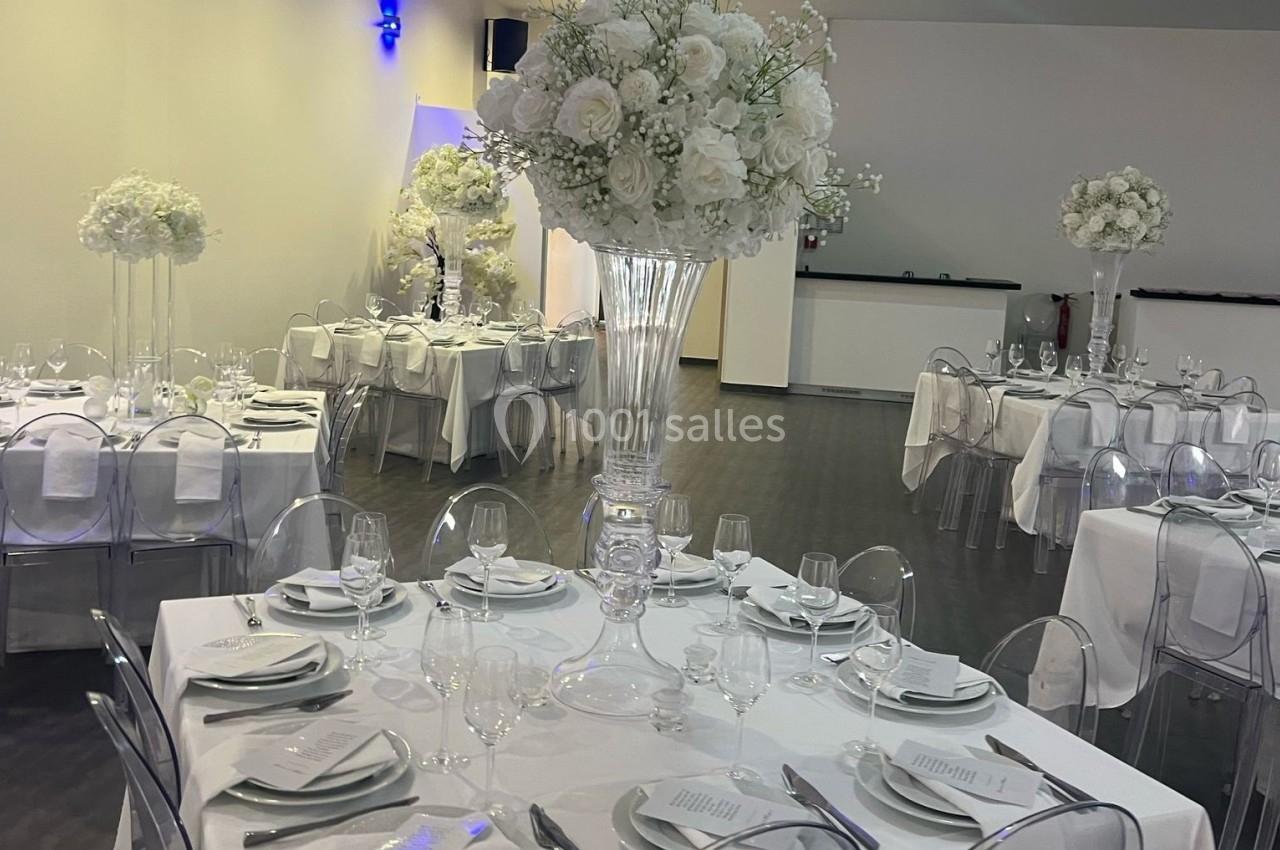 Salle de réception décorée avec des tables élégantes, nappes blanches, chaises transparentes et centres de table fleuris.