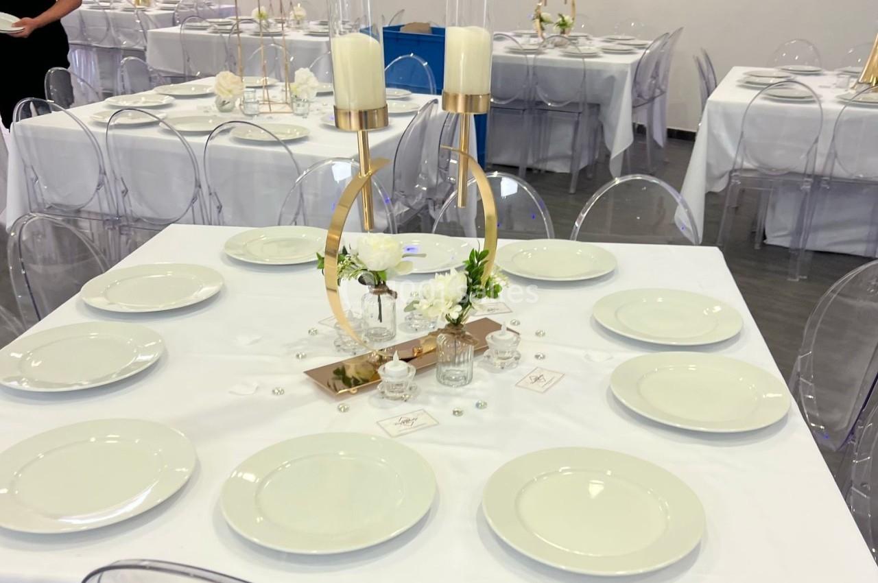 Table dressée avec une nappe blanche, vaisselle sobre, centre de table doré avec bougies et fleurs blanches.
