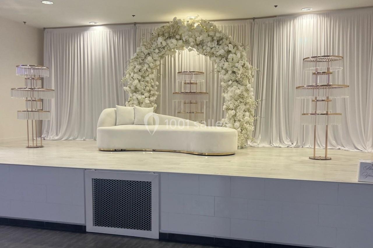Canapé blanc devant une arche florale ornée de fleurs blanches, entouré de présentoirs métalliques sur une scène.