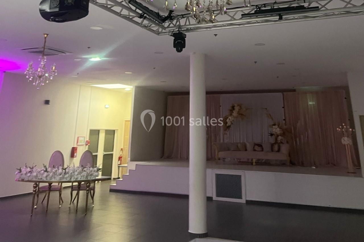 Salle de réception avec scène décorée de rideaux beiges et fleurs, table et chaises élégantes au premier plan.