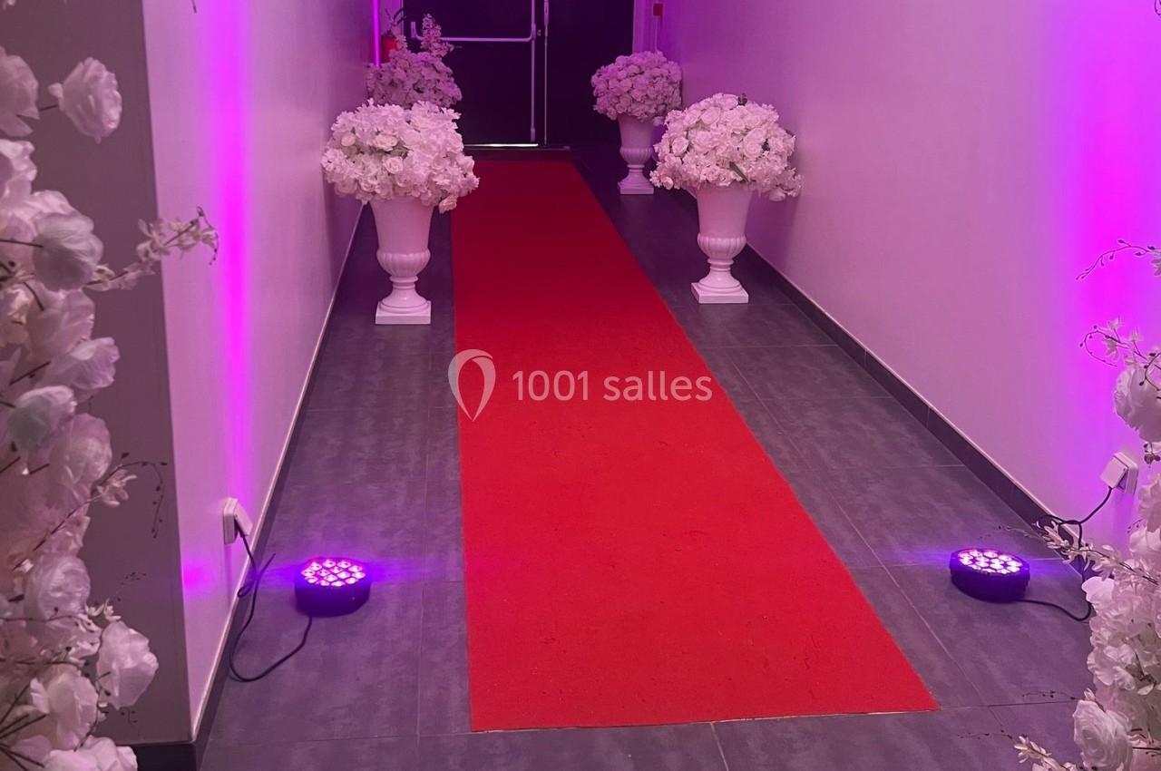 Allée avec tapis rouge bordée de vases blancs remplis de fleurs roses, éclairée par des lumières violettes.