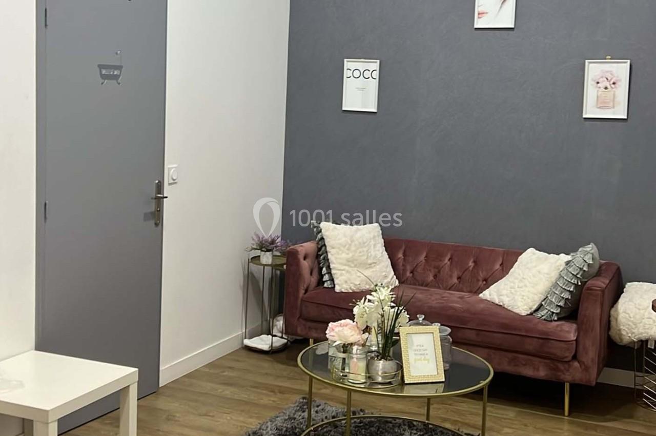 Salon moderne avec canapé rose, table basse en verre, décorations florales et cadres accrochés sur un mur gris.