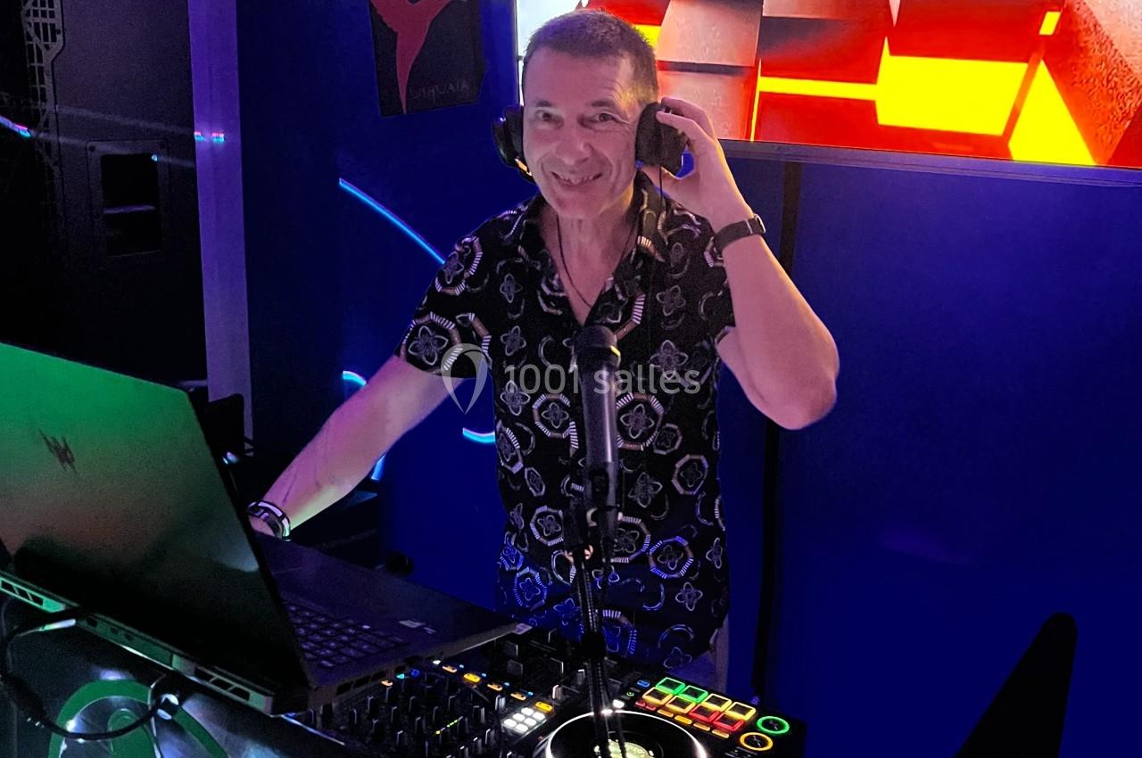 Un DJ souriant mixe de la musique dans un espace sombre, éclairé par des lumières colorées et un écran lumineux.