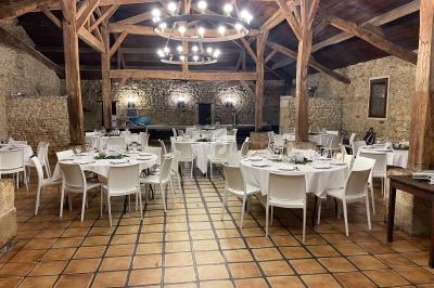 Salle de réception rustique avec tables rondes dressées, chaises blanches et poutres apparentes en bois.