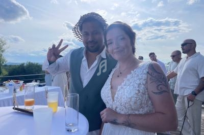 Un couple souriant en tenue de mariage pose sur une terrasse ensoleillée avec des invités en arrière-plan.