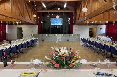 Salle décorée pour un événement avec tables dressées, fleurs au premier plan et scène avec écran en arrière-plan.
