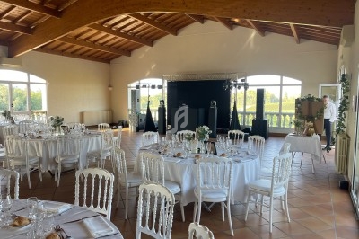 Salle de réception lumineuse avec tables rondes dressées, chaises blanches et vue sur un paysage verdoyant.