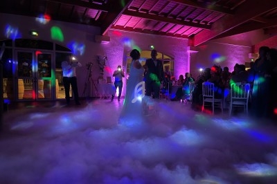 Un couple danse au milieu d'une piste enveloppée de fumée, éclairée par des lumières colorées dans une salle festive.