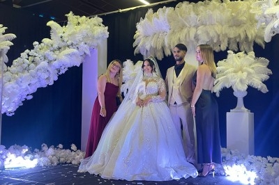 Un couple en tenue de mariage pose avec deux invités devant une décoration florale et des plumes blanches.