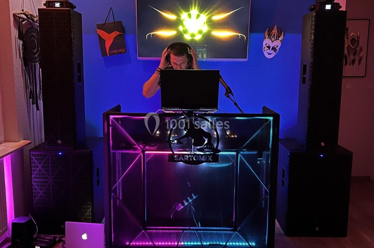 Un DJ mixe de la musique derrière une table lumineuse entourée de haut-parleurs dans une pièce décorée.