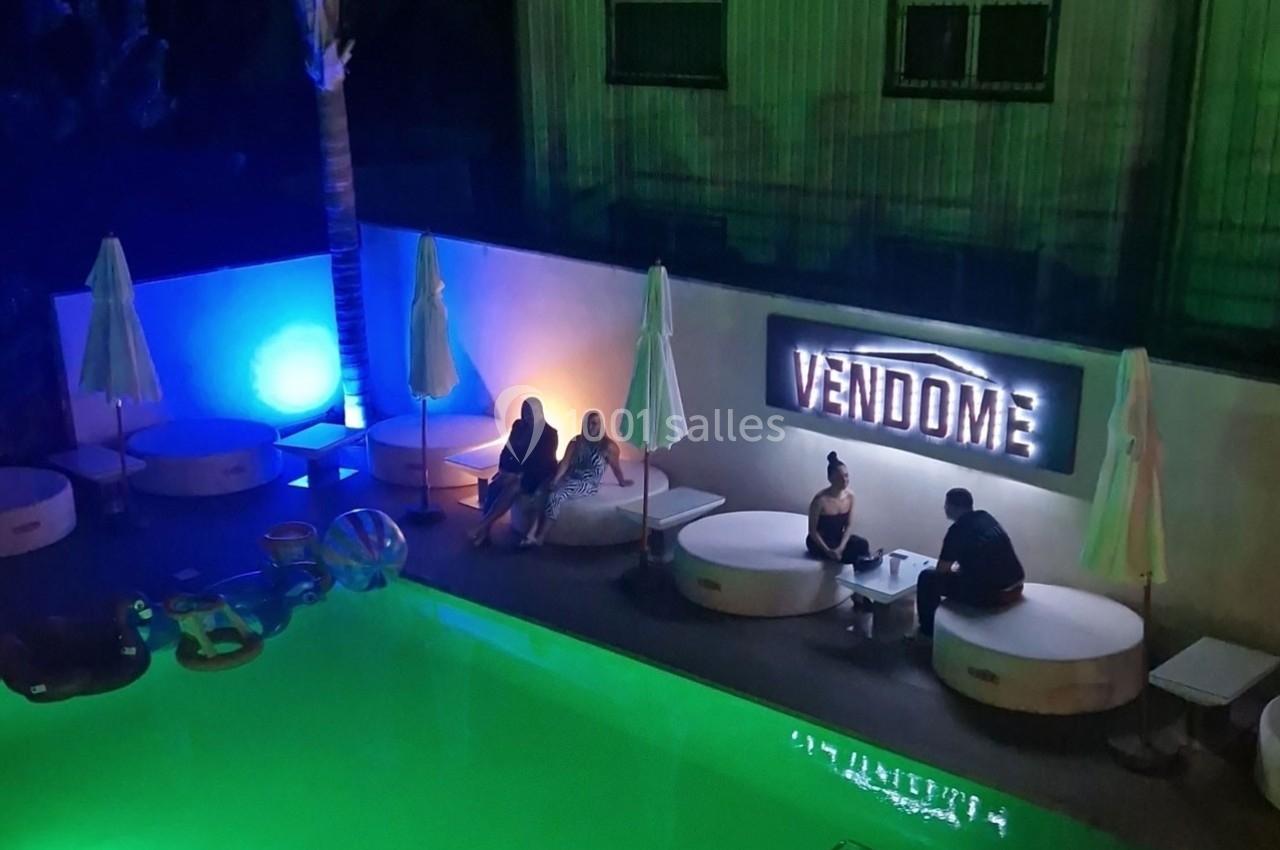 Personnes assises près d'une piscine éclairée, entourées de parasols fermés, avec un panneau lumineux ’Vendôme’.