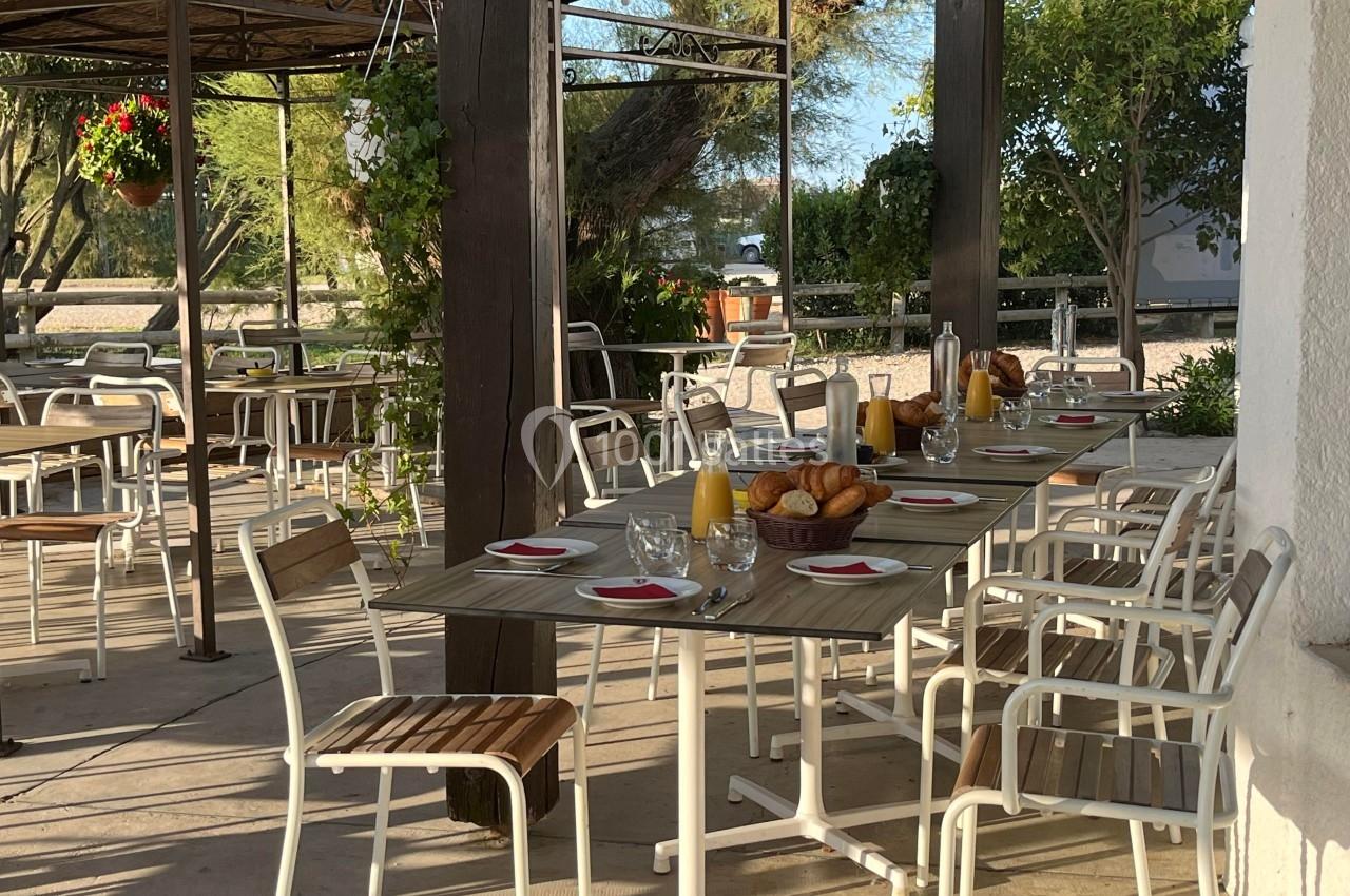 Terrasse ombragée avec tables dressées pour le petit-déjeuner, contenant croissants, jus d'orange et vaisselle.