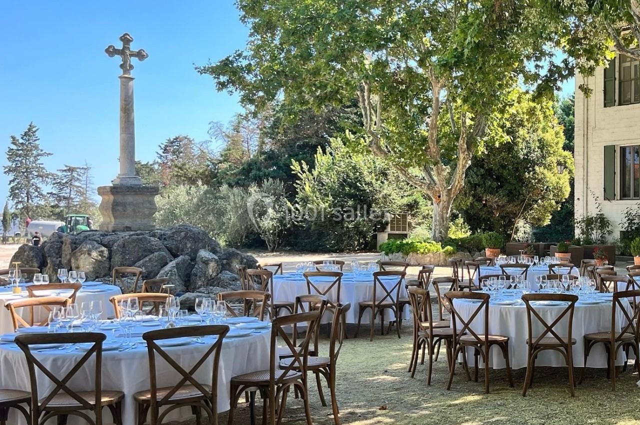 Tables rondes dressées avec nappes blanches et chaises en bois, disposées en extérieur sous des arbres près d'une croix en…