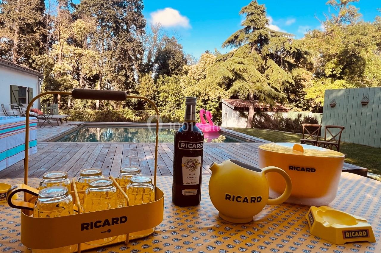 Table avec des verres, une bouteille et des accessoires Ricard, près d'une piscine entourée de verdure.