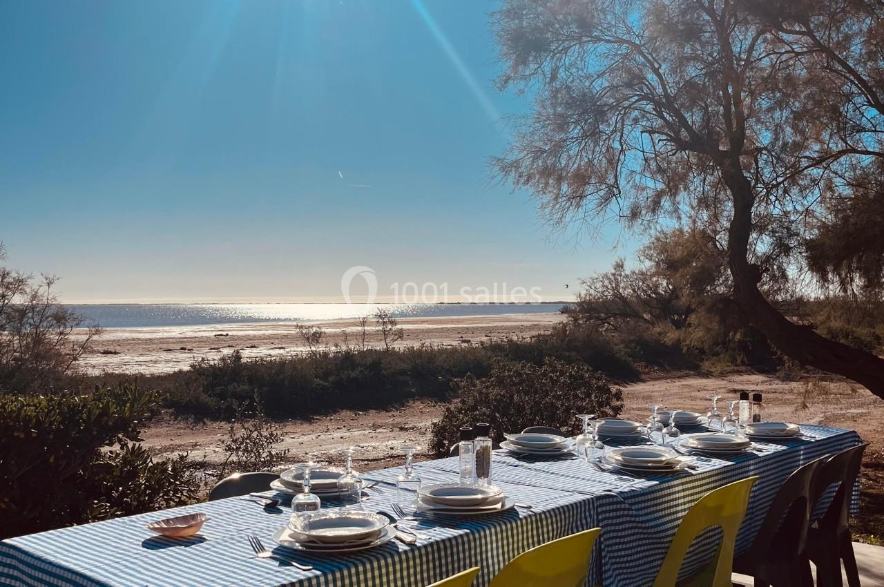 Table dressée avec nappe à carreaux bleus, chaises colorées, vue sur une plage ensoleillée et un arbre en arrière-plan.