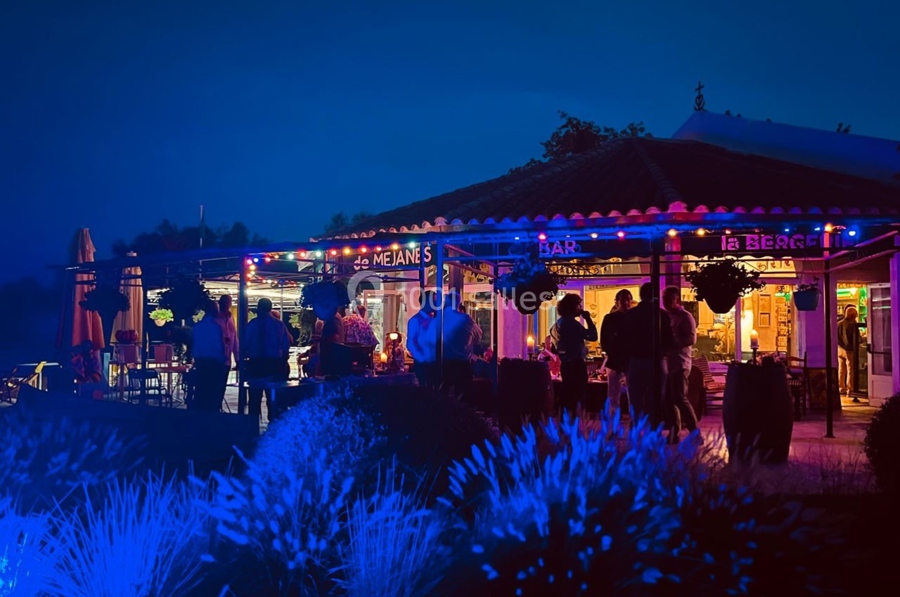 Terrasse d'un bar-restaurant éclairée par des guirlandes lumineuses, avec des clients rassemblés en soirée.