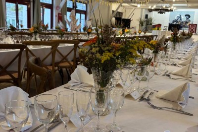 Tables élégamment dressées avec nappes blanches, serviettes pliées, verres et décorations florales dans une salle lumineuse.