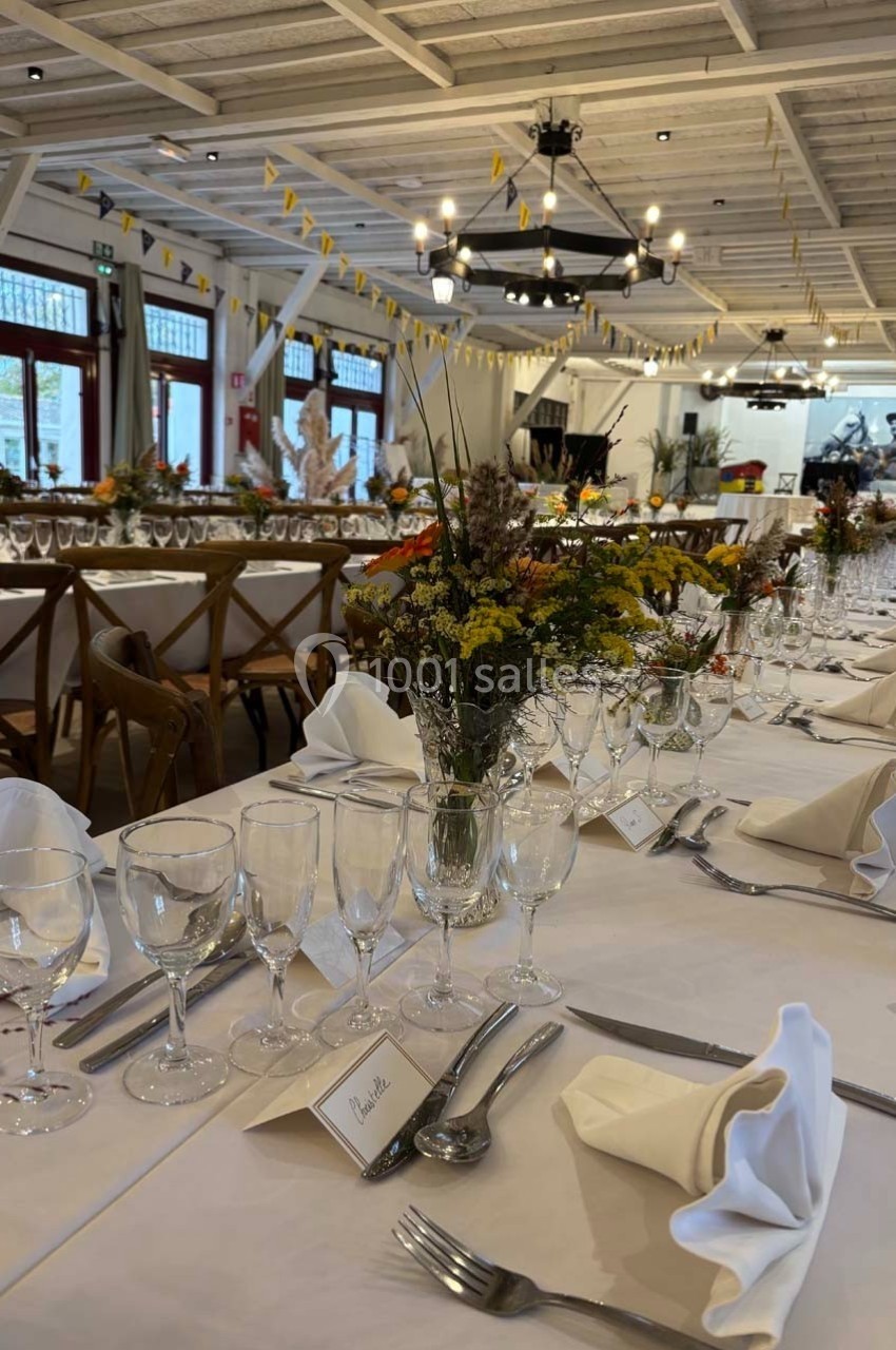 Table dressée pour un événement avec nappes blanches, verres, couverts, serviettes pliées et décorations florales.