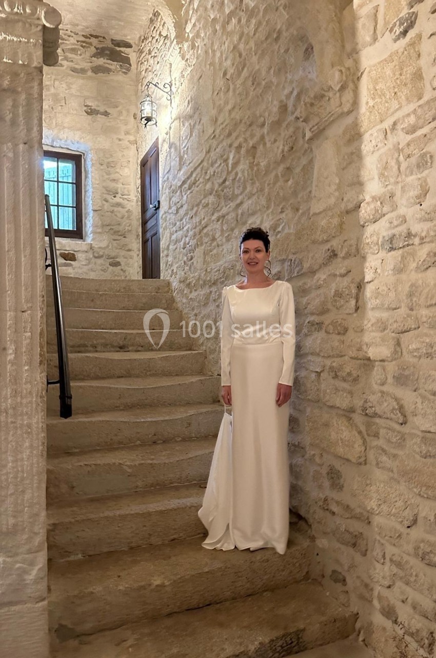 Une femme en robe blanche se tient près d'un escalier en pierre dans un intérieur ancien à murs de pierre apparente.