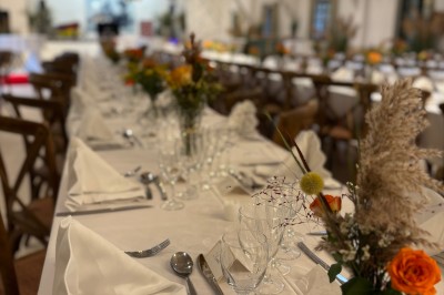 Tables élégamment dressées avec nappes blanches, serviettes pliées, verres et décorations florales dans une salle lumineuse.
