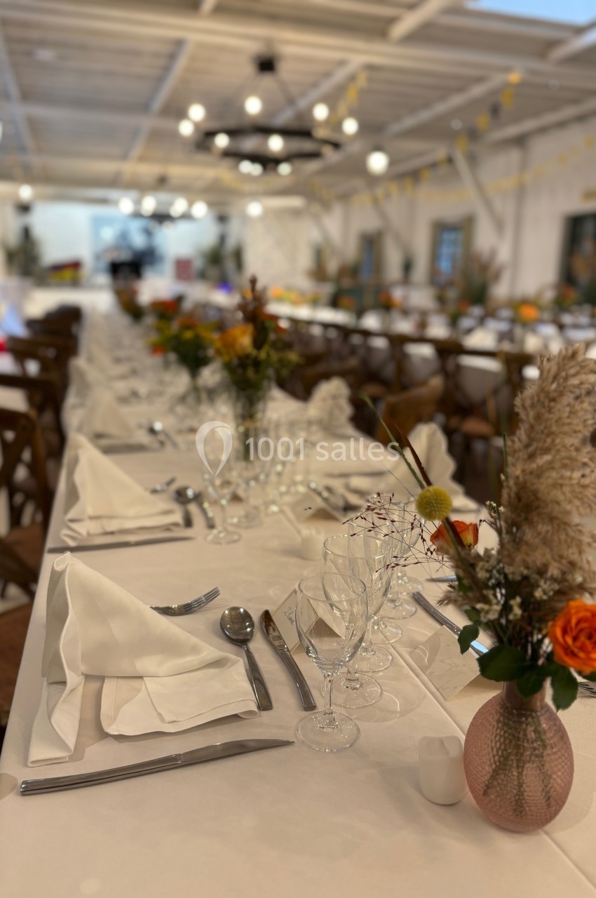 Tables élégamment dressées avec nappes blanches, serviettes pliées, verres et décorations florales dans une salle lumineuse.