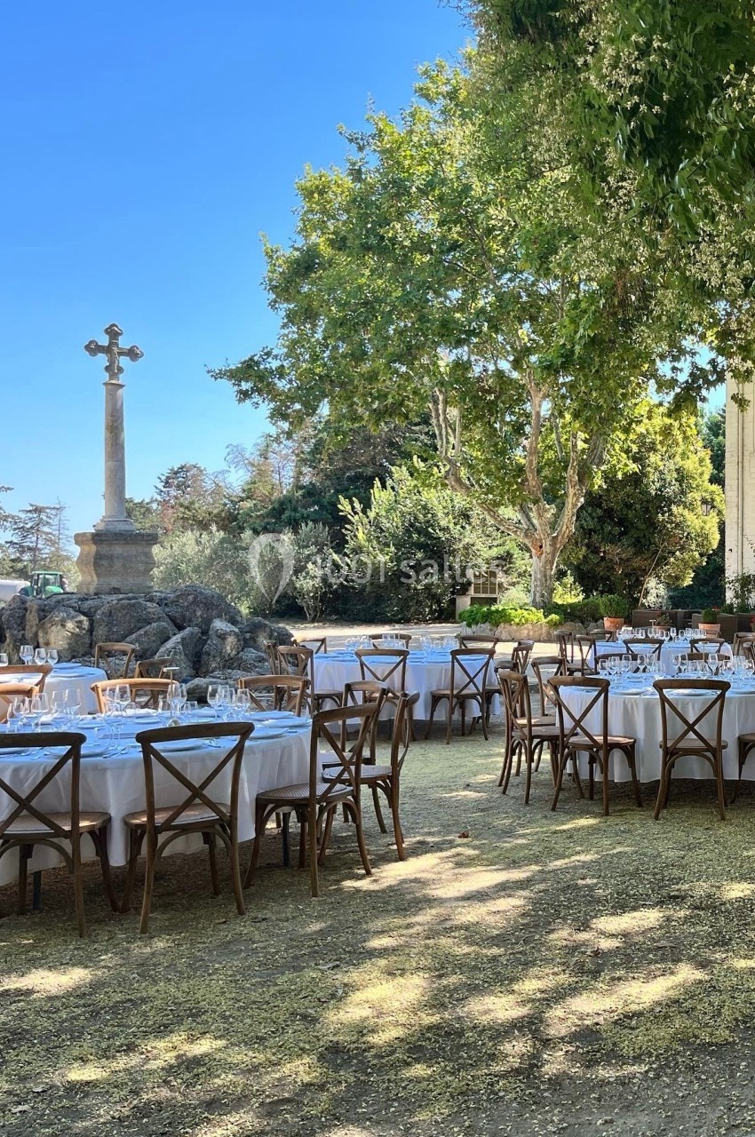 Tables rondes dressées avec nappes blanches et chaises en bois, disposées en extérieur sous des arbres près d'une croix en…