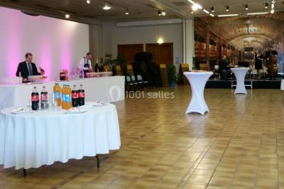 Salle de réception décorée avec des tables rondes, nappes blanches, rubans rouges et tonneaux en arrière-plan.