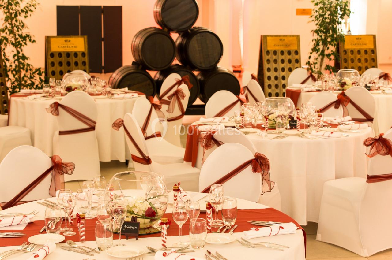 Salle de réception décorée avec des tables rondes, nappes blanches, rubans rouges et tonneaux en arrière-plan.