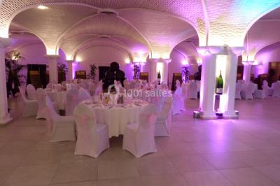Salle de réception décorée avec des tables rondes, nappes blanches, rubans rouges et tonneaux en arrière-plan.