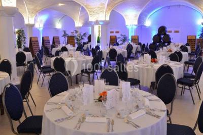 Salle de réception décorée avec des tables rondes, nappes blanches, rubans rouges et tonneaux en arrière-plan.