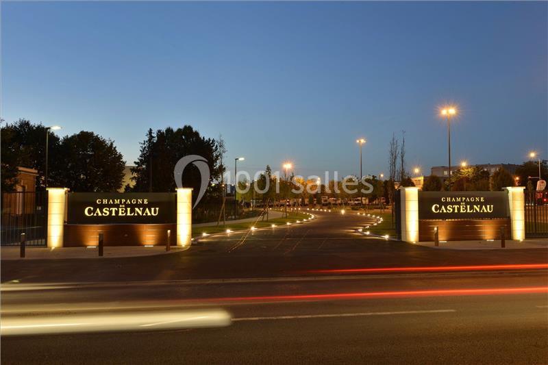 Entrée illuminée de nuit avec deux panneaux indiquant ’Champagne Castelnau’, entourée d'arbres et de lampadaires.