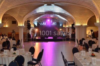 Salle de réception décorée avec des tables rondes, nappes blanches, rubans rouges et tonneaux en arrière-plan.