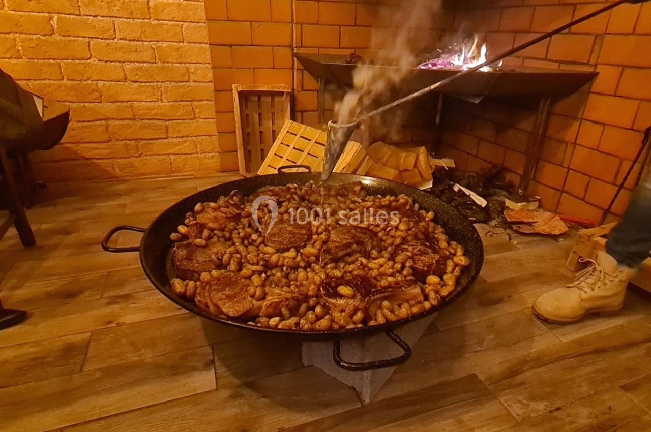 Grande poêle contenant des morceaux de viande et des pommes de terre, cuite sur un feu dans un cadre rustique.