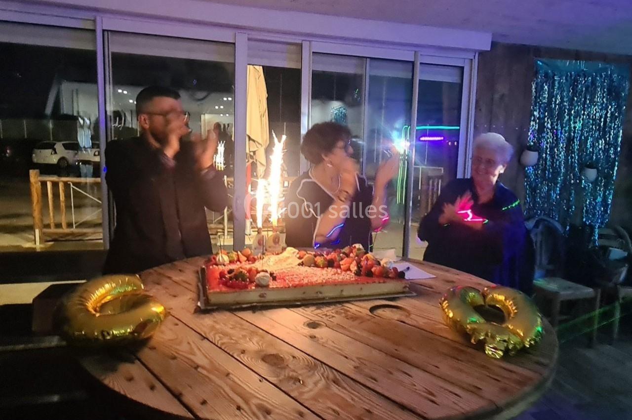 Trois personnes applaudissent autour d'un gâteau d'anniversaire décoré, avec des bougies allumées sur une table en bois.
