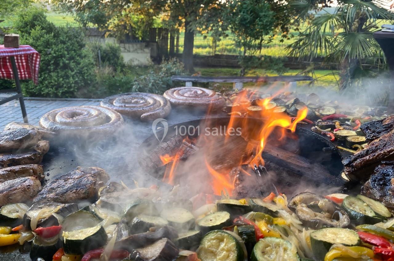 Barbecue en plein air avec légumes grillés, saucisses en spirale et flammes vives sur une plancha, entouré de verdure.
