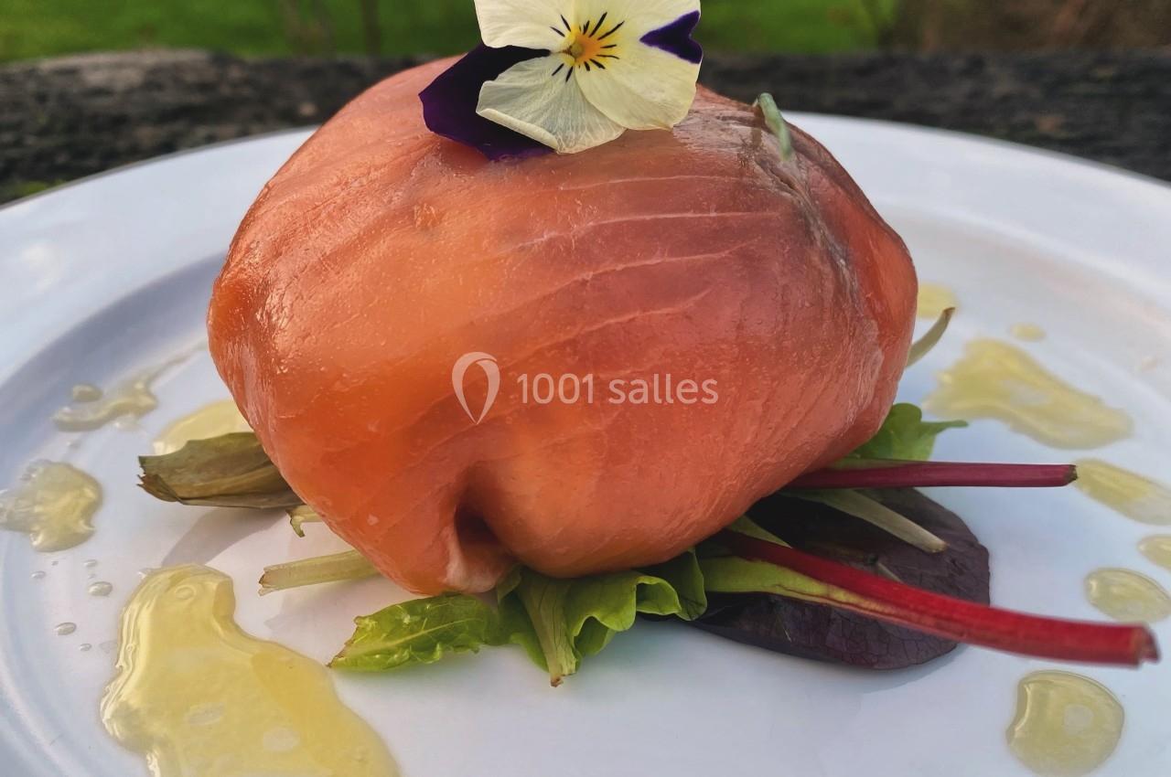 Ballotine de saumon fumé garnie de jeunes pousses, décorée d'une fleur comestible, servie avec un filet d'huile.