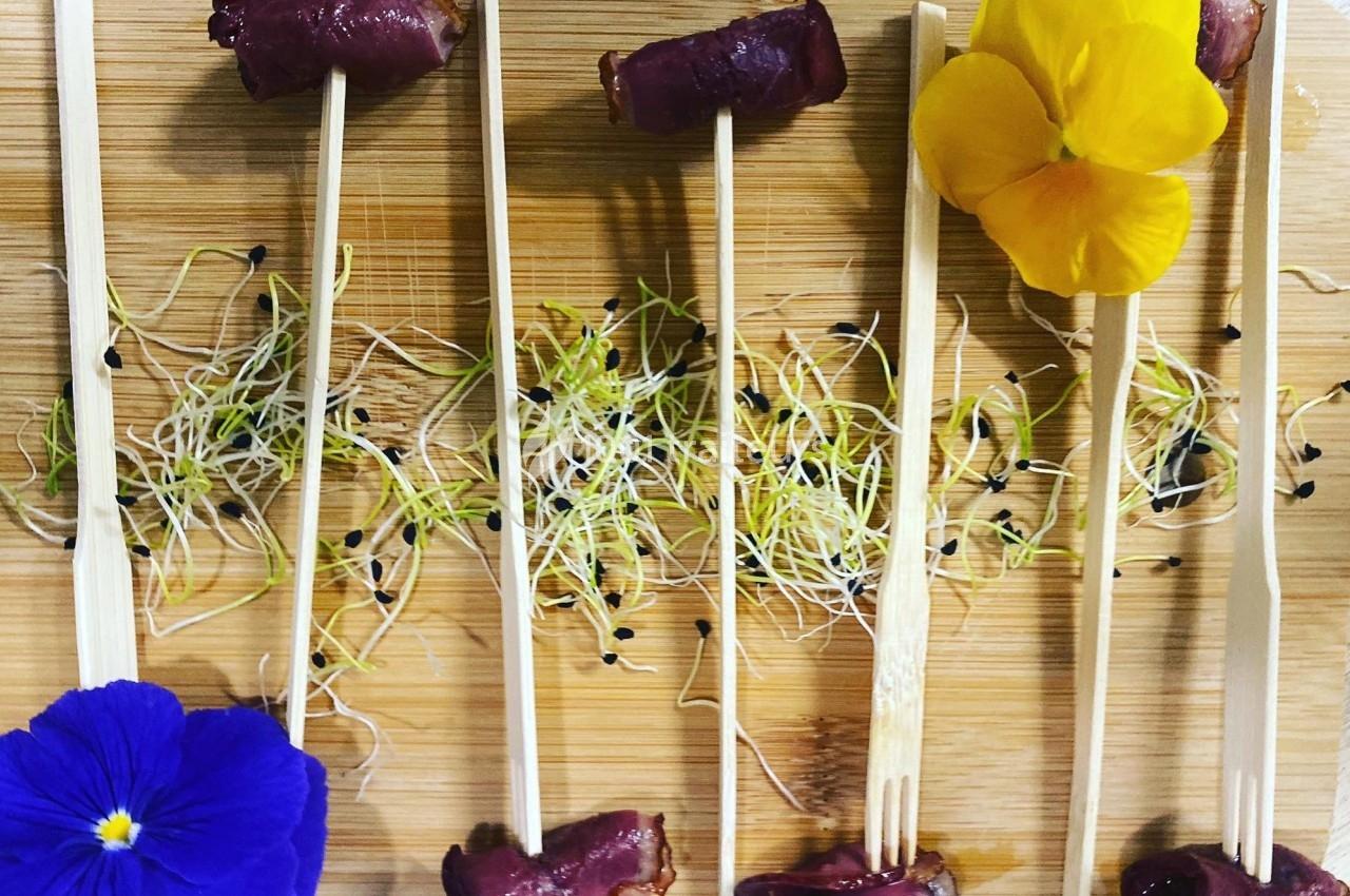 Bouchées de viande sur des piques en bois, décorées de fleurs comestibles et de pousses sur une planche en bois.
