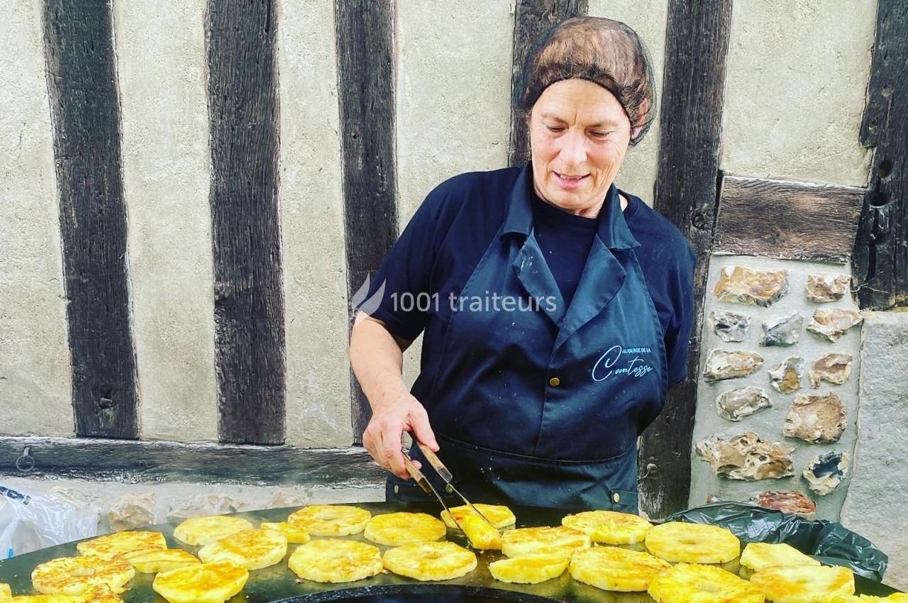 Femme en tablier noir cuisant des tranches de légumes dorés sur une grande plaque chauffante devant un mur à colombages.