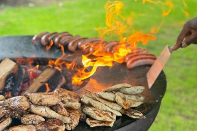 Deux hommes cuisinent des aliments sur un grand barbecue en plein air, éclairé par les flammes, à la tombée de la nuit.