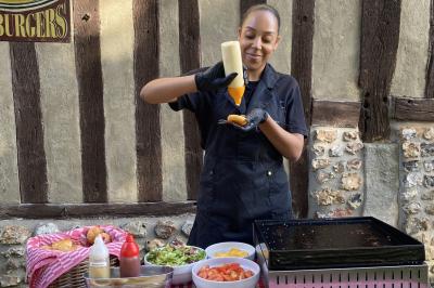 Deux hommes cuisinent des aliments sur un grand barbecue en plein air, éclairé par les flammes, à la tombée de la nuit.
