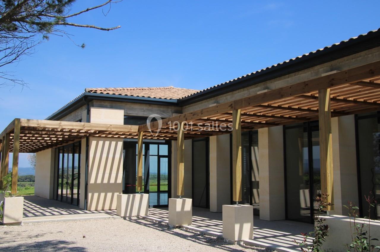 Façade d'une maison moderne avec pergola en bois, grandes baies vitrées et toit en tuiles, sous un ciel bleu.