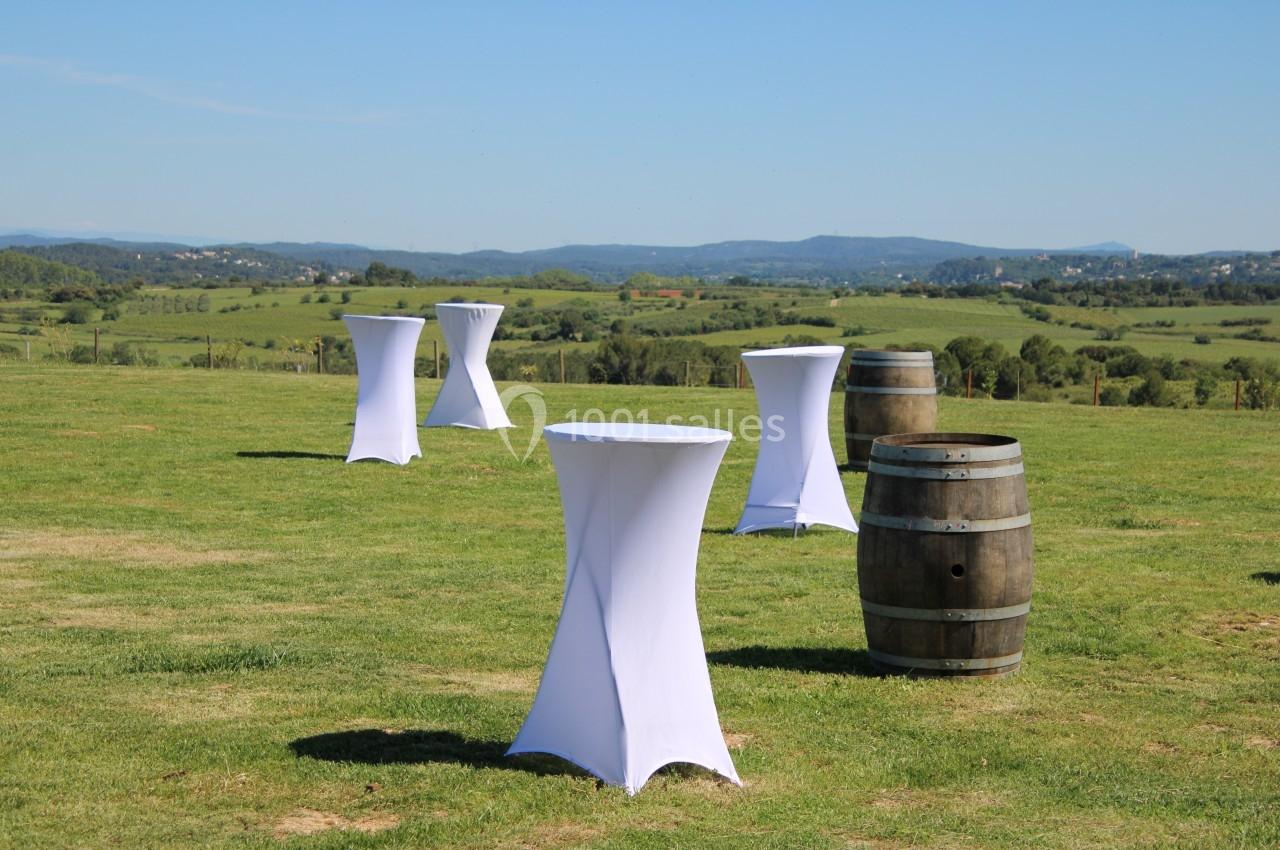 Tables hautes blanches et tonneau en bois disposés sur une pelouse avec vue sur un paysage vallonné.