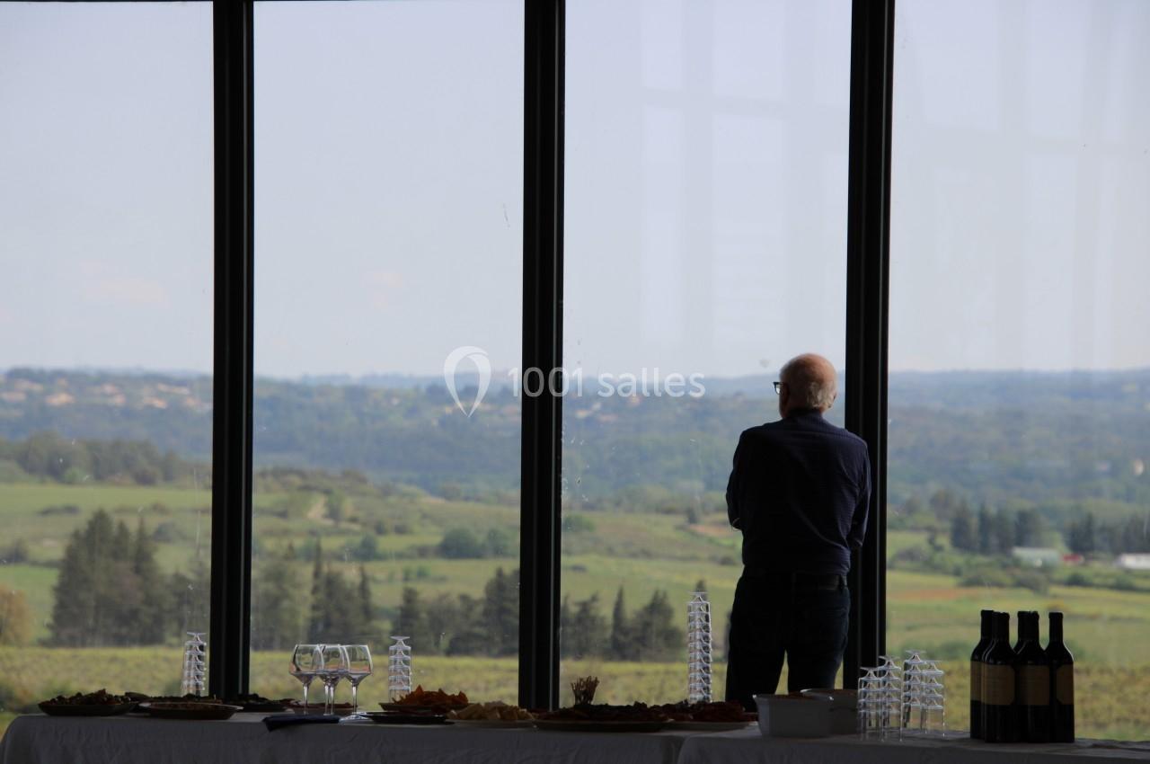 Un homme regarde un paysage vallonné à travers de grandes baies vitrées, avec une table dressée au premier plan.