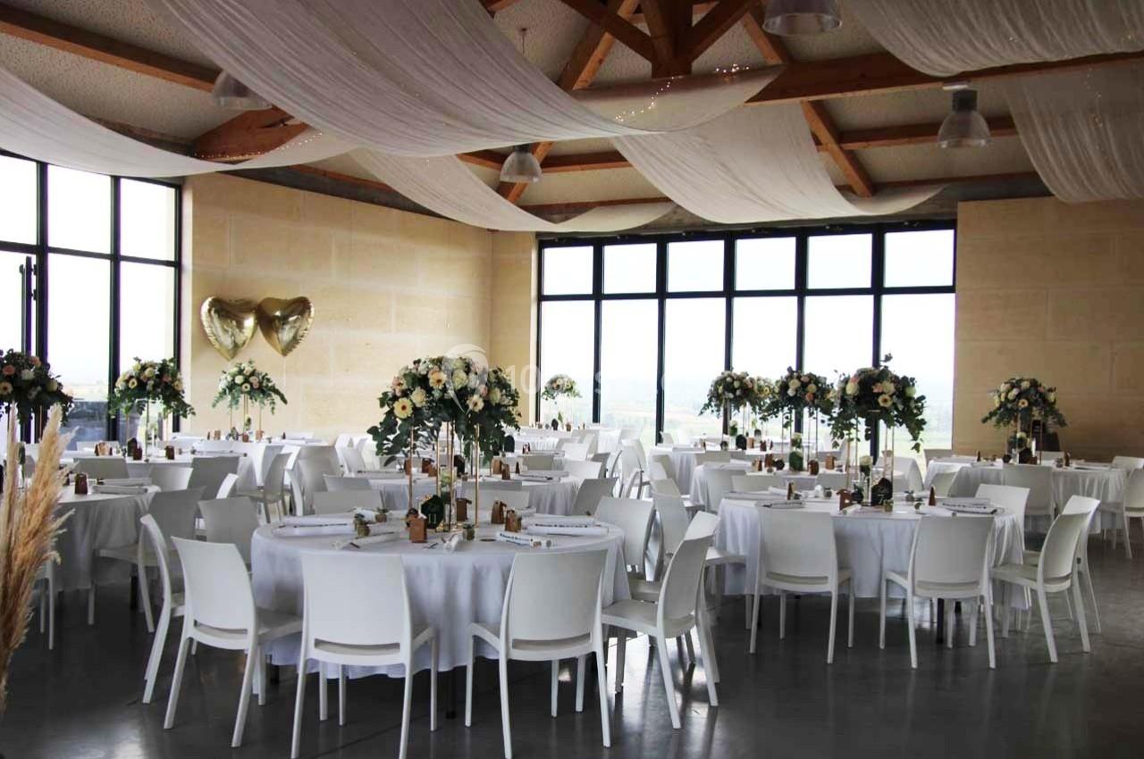Salle de réception décorée avec des tables rondes, des chaises blanches, des fleurs et des ballons dorés en forme de cœur.
