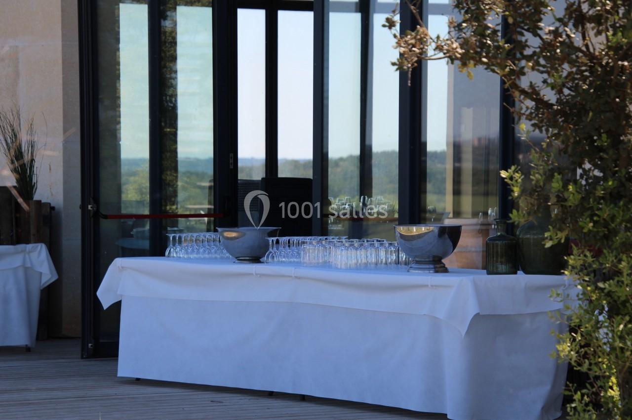 Table dressée en extérieur avec nappes blanches, verres alignés et bols, devant des baies vitrées donnant sur un paysage.