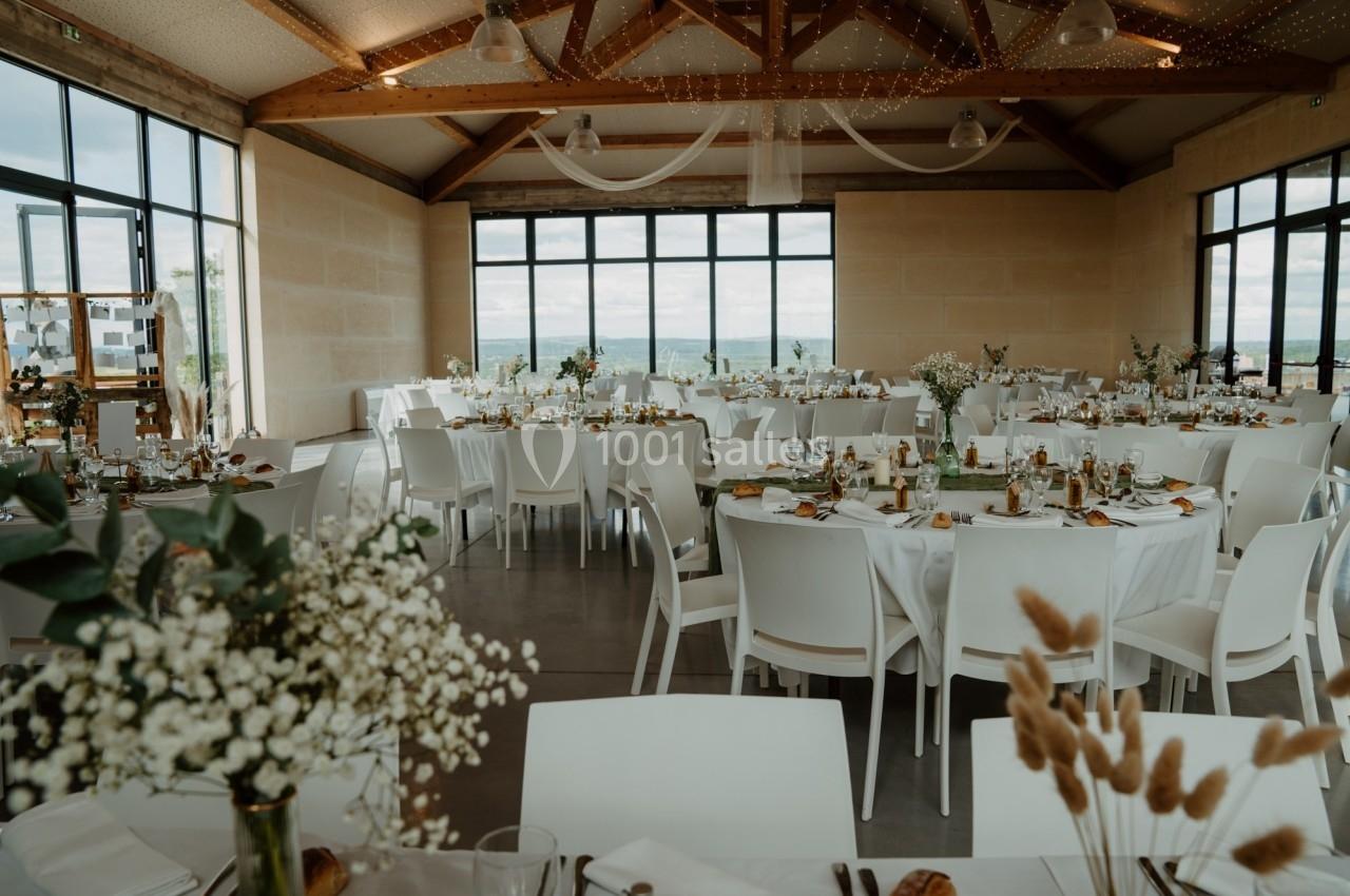 Salle de réception lumineuse avec tables décorées de fleurs et couverts, offrant une vue sur l'extérieur par de grandes…