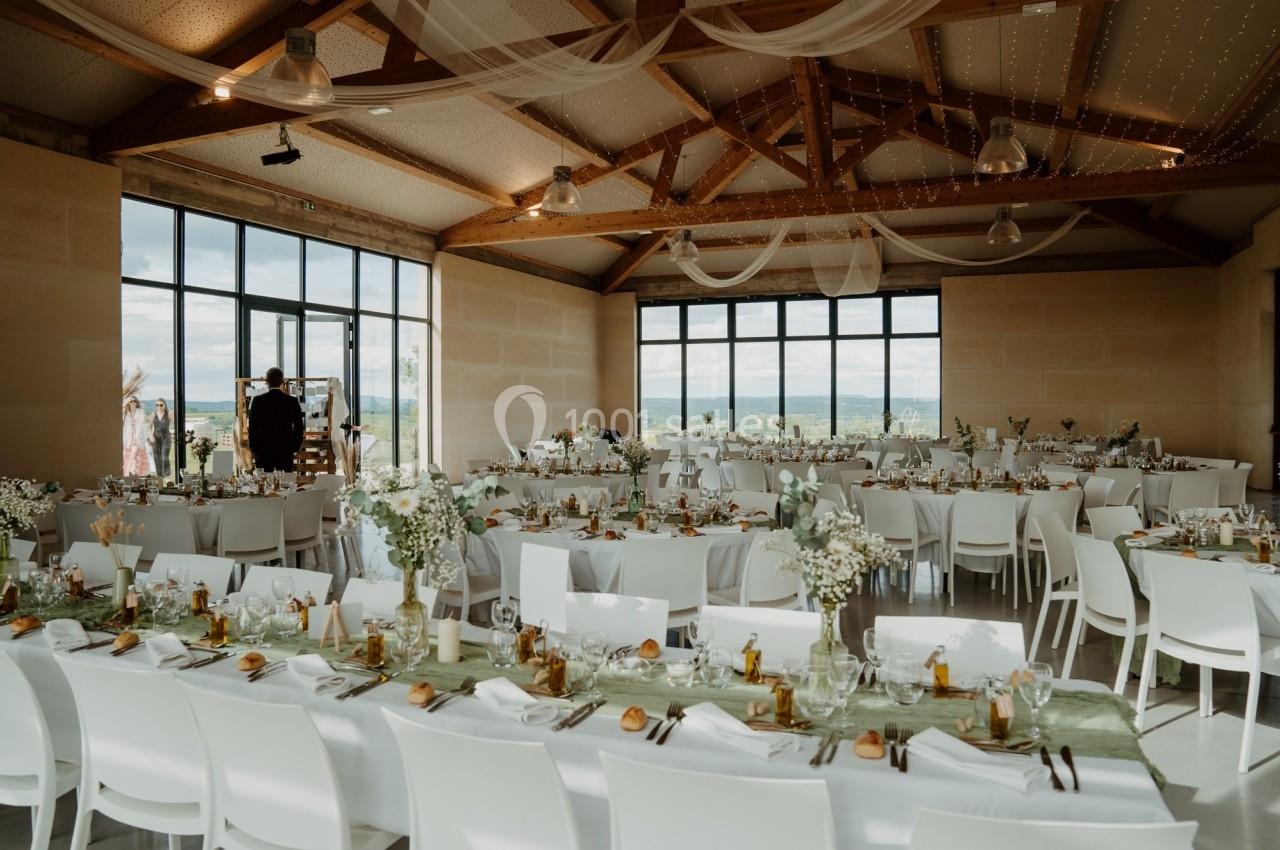 Salle de réception lumineuse décorée pour un mariage, avec tables dressées, chaises blanches et grandes baies vitrées.
